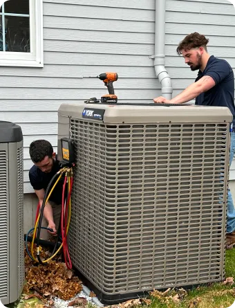 Two technicians service an outdoor HVAC unit next to a house; one checks wires while the other stands nearby with a tool.