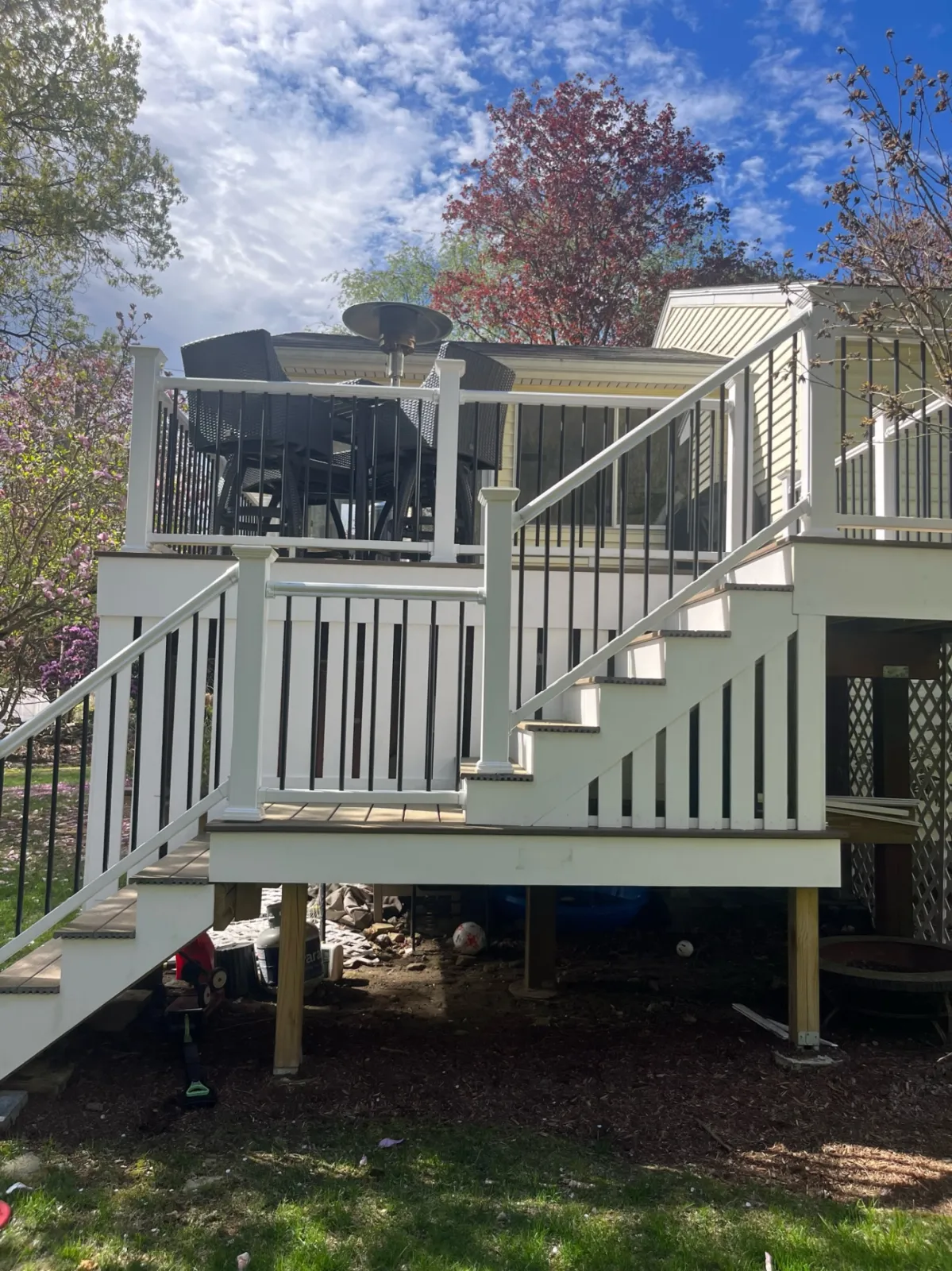 Outdoor deck with stairs, patio furniture, and umbrella heater, surrounded by trees under blue sky with clouds.