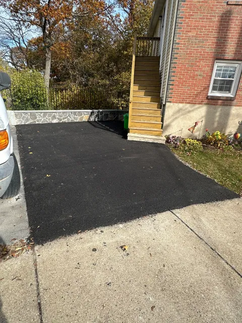 Black asphalt driveway beside a brick building, next to a parked silver car and stacked wooden pallets