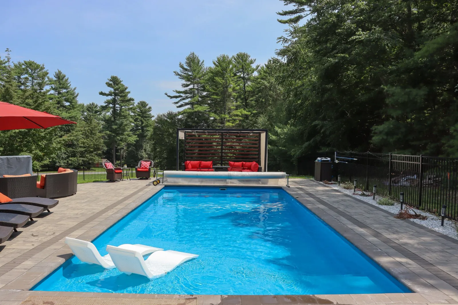 Swimming pool with two white lounge chairs, surrounded by outdoor furniture including chairs, sofas, and a red umbrella, with trees in the background.