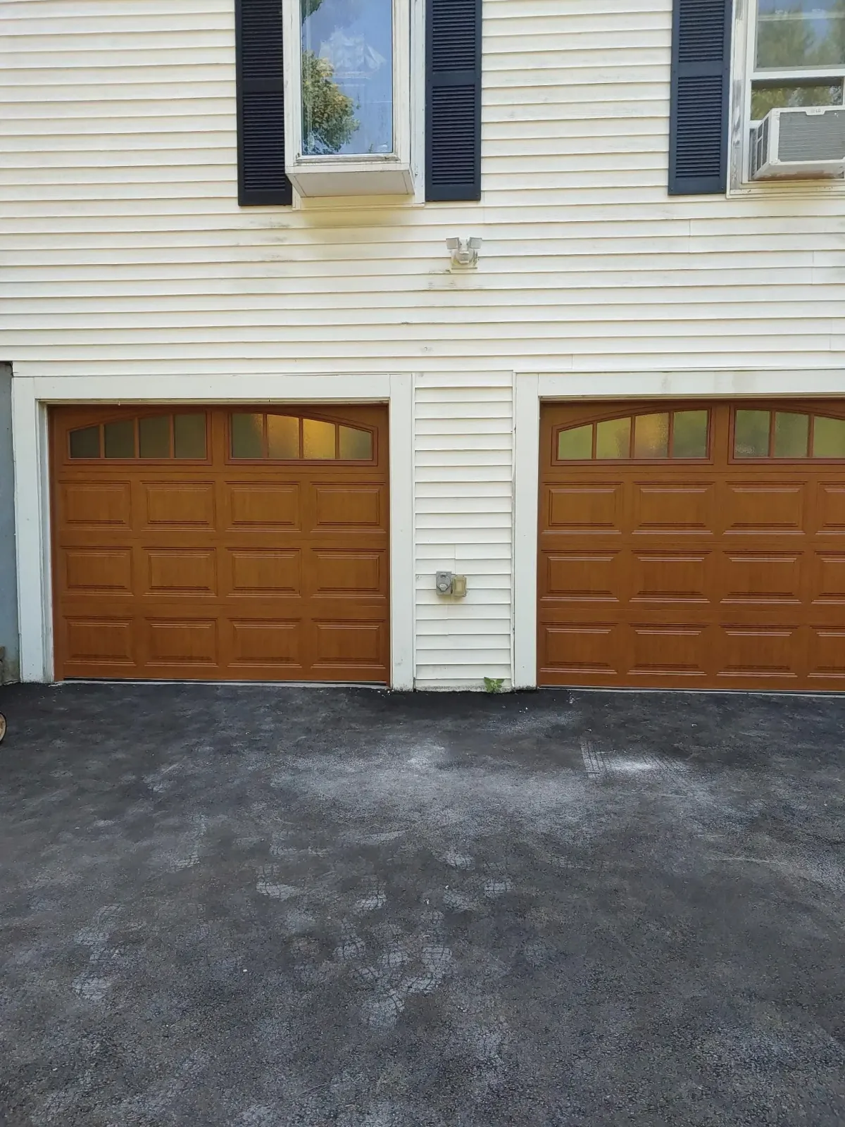 Two brown garage doors on a weathered white house. Black asphalt driveway.