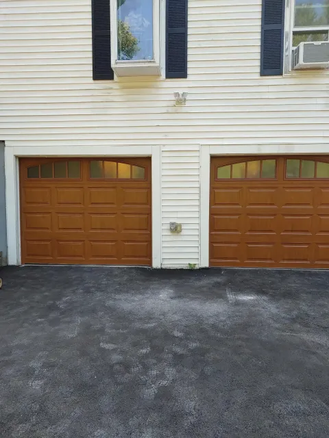 Two brown garage doors on a weathered white house. Black asphalt driveway.