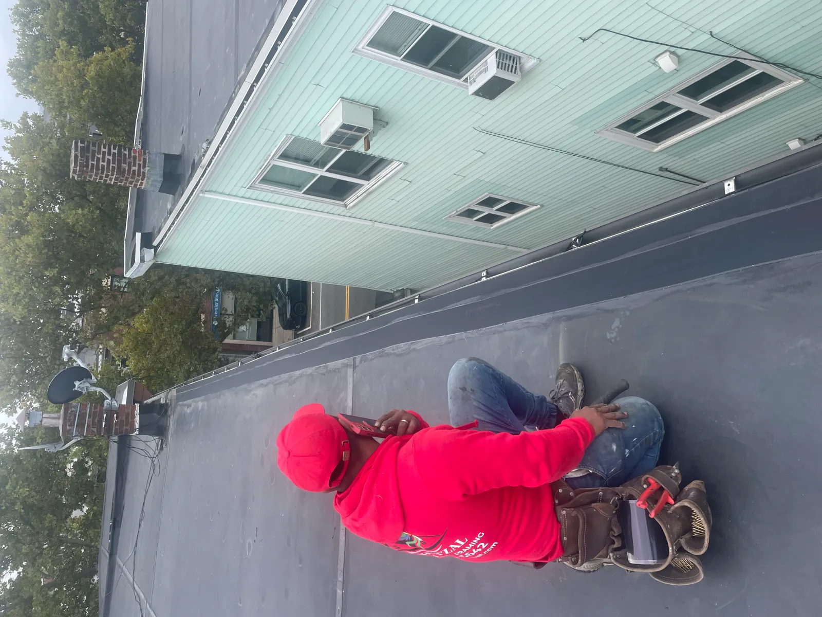 Person in red hoodie sitting on a flat roof, looking at a nearby building; tools and a chimney visible.