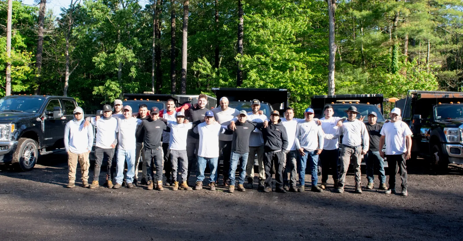 A group of 19 men standing outdoors in front of black trucks, posing for a group photo with trees in the background.