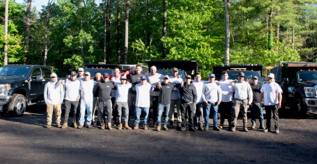 A group of 19 men standing outdoors in front of black trucks, posing for a group photo with trees in the background.
