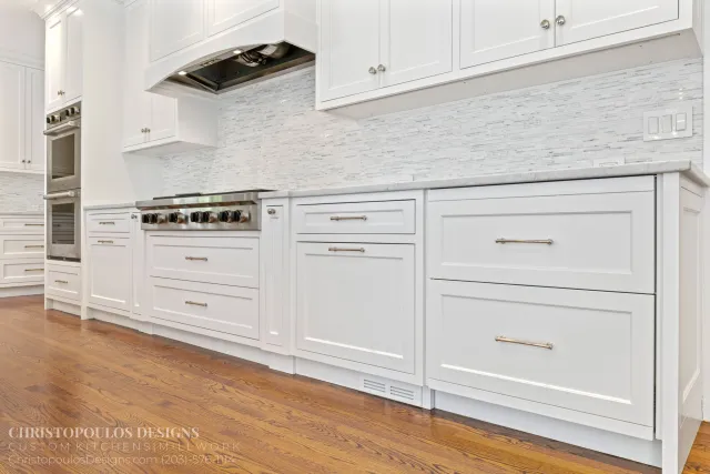Modern white kitchen with white cabinets, marble countertops, and a glass tile backsplash, hardwood flooring, and built-in appliances.