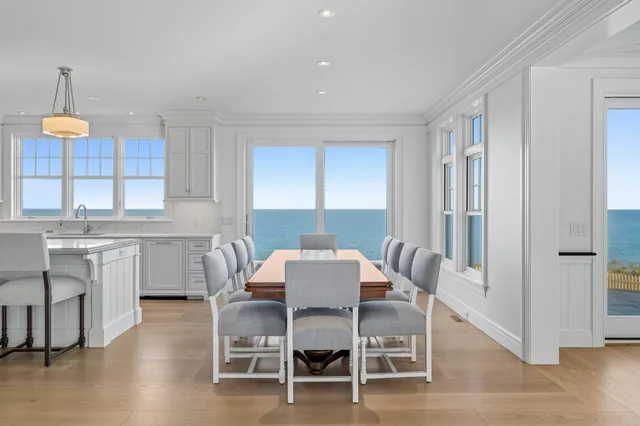 Bright dining room interior of the Luscombe Lane project in Dennis, MA, featuring a wood dining table, gray upholstered chairs, shiplap walls, and large windows with ocean views