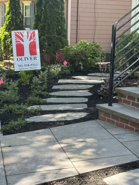 Stone pathway with stepping stones leading to a house entrance with brick steps and black railing, surrounded by garden plants and a landscaping company sign.