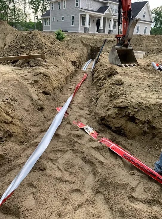 Excavator digging a trench beside a house, with exposed white and red utility pipes in sandy soil