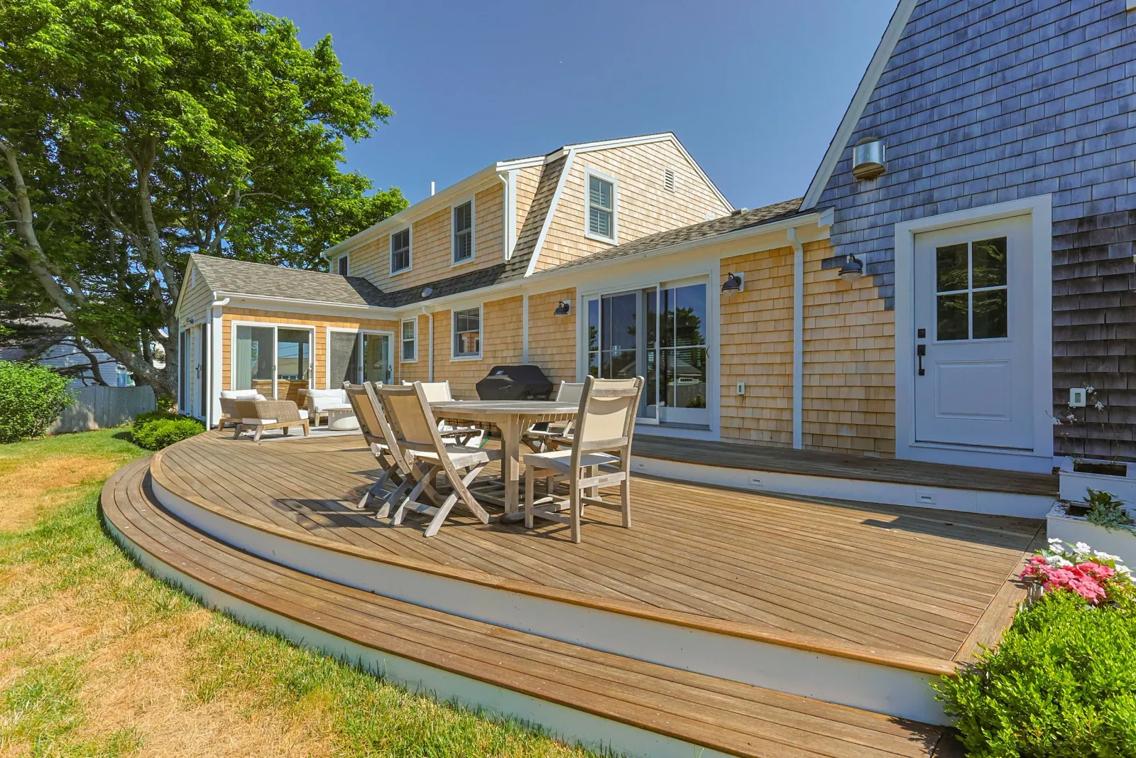 Backyard deck with outdoor furniture, a grill, and sliding glass door, adjacent to a yellow and blue house with a tree on the left and a flower bed on the right. Custom deck and exterior enhancements by Watkins Works