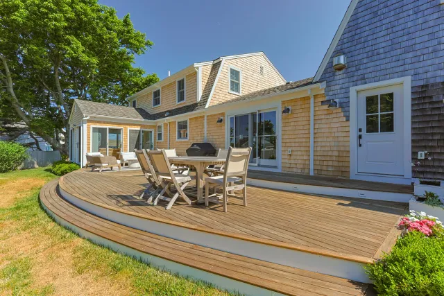Backyard deck with outdoor furniture, a grill, and sliding glass door, adjacent to a yellow and blue house with a tree on the left and a flower bed on the right. Custom deck and exterior enhancements by Watkins Works