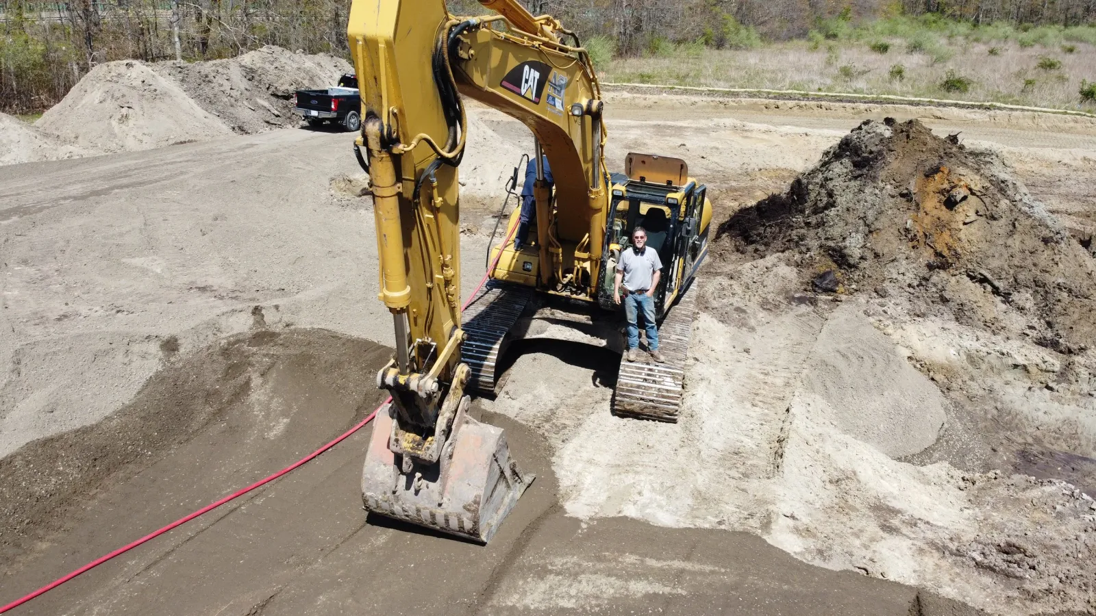 A construction site featuring a yellow Caterpillar excavator with a person standing beneath its arm. The site has dirt and uneven ground with mounds of soil, and a black pickup truck is visible in the background in a rural area with trees.
