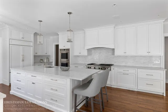 Modern white kitchen with large island, pendant lights, built-in appliances, and hardwood floors.