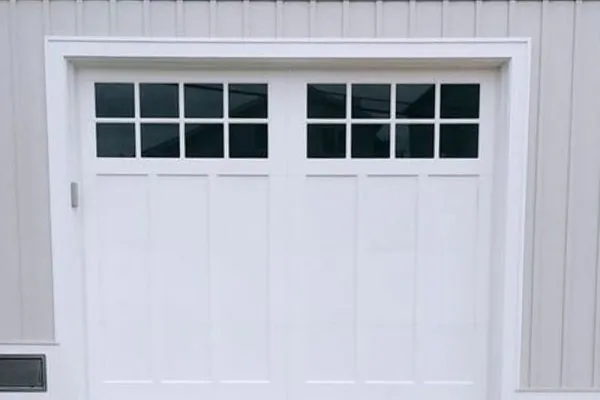 White garage door with grid windows, showcasing a recent garage door installation. The door is framed by light gray siding and gravel, creating a clean, modern aesthetic.