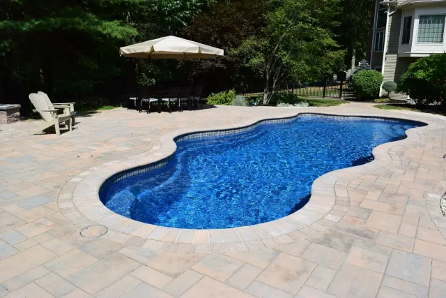 Swimming pool with blue water surrounded by beige paving stones. A patio umbrella and a chair are visible.
