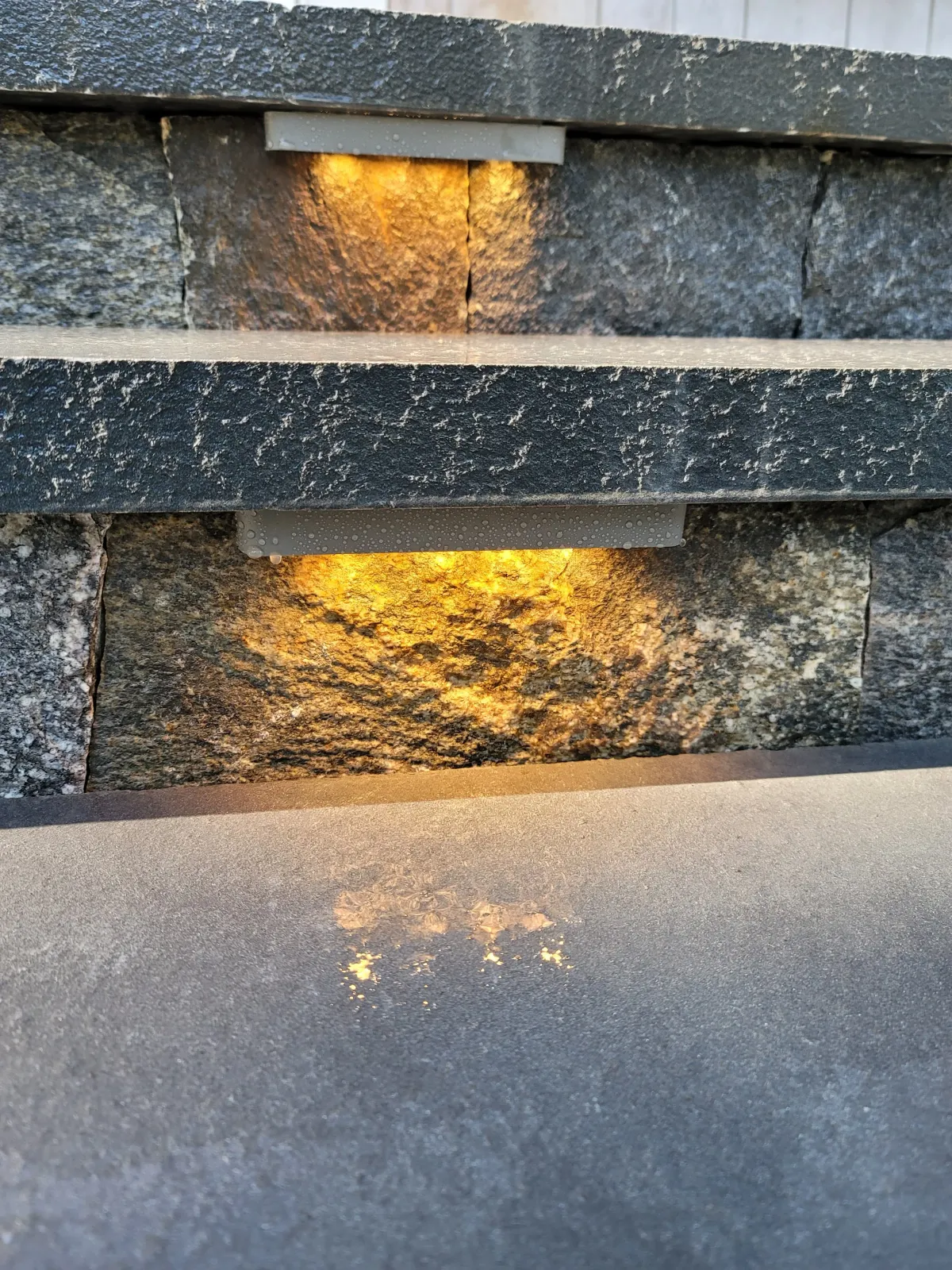 Close-up view of a section of a stone and metal outdoor structure, with small lights casting a warm glow onto the stones and reflecting on the surface below.