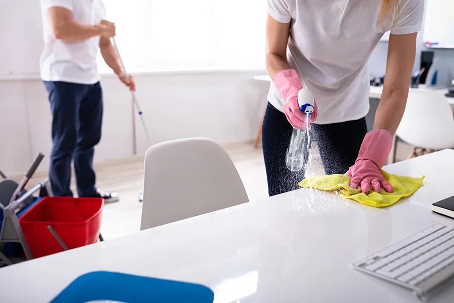 Two people cleaning an office highlight the difference in tasks: one sprays a desk while the other mops the floor in the background.