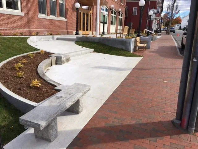 Sidewalk with a curved concrete path, brick-paved street, benches, street lamps, and a brick building in a small town setting.