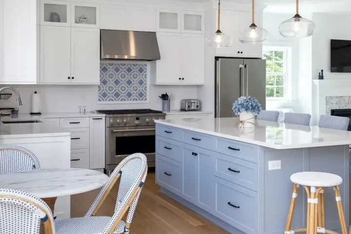 Bright white and light blue kitchen with a large island, stainless steel appliances, glass pendant lights, and a blue hydrangea centerpiece. Woven chairs nearby.
