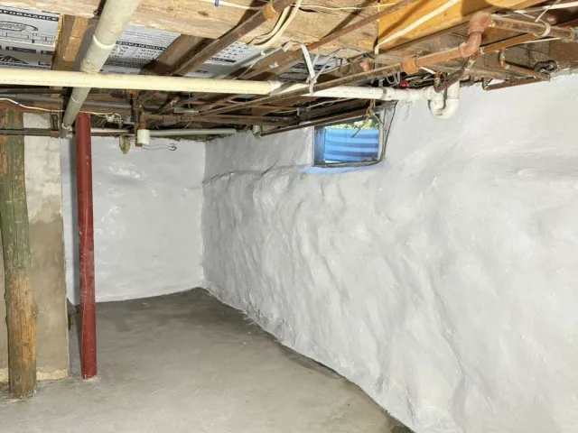 Unfinished basement crawlspace with white insulated walls, exposed pipes and joists, and a small blue window.