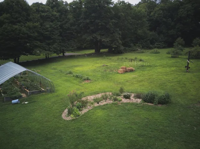 A backyard with a small garden bed, a greenhouse, hay bales, colorful plants, and trees in the background.