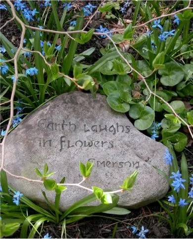 Stone with "Earth laughs in flowers - Emerson" engraved, surrounded by grass and small blue flowers.