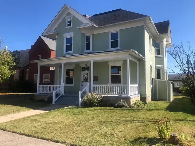 A two-story light green house with a large front porch and white railing, black roof, surrounded by a lawn with a sidewalk leading to the front steps, and other houses visible in the background.