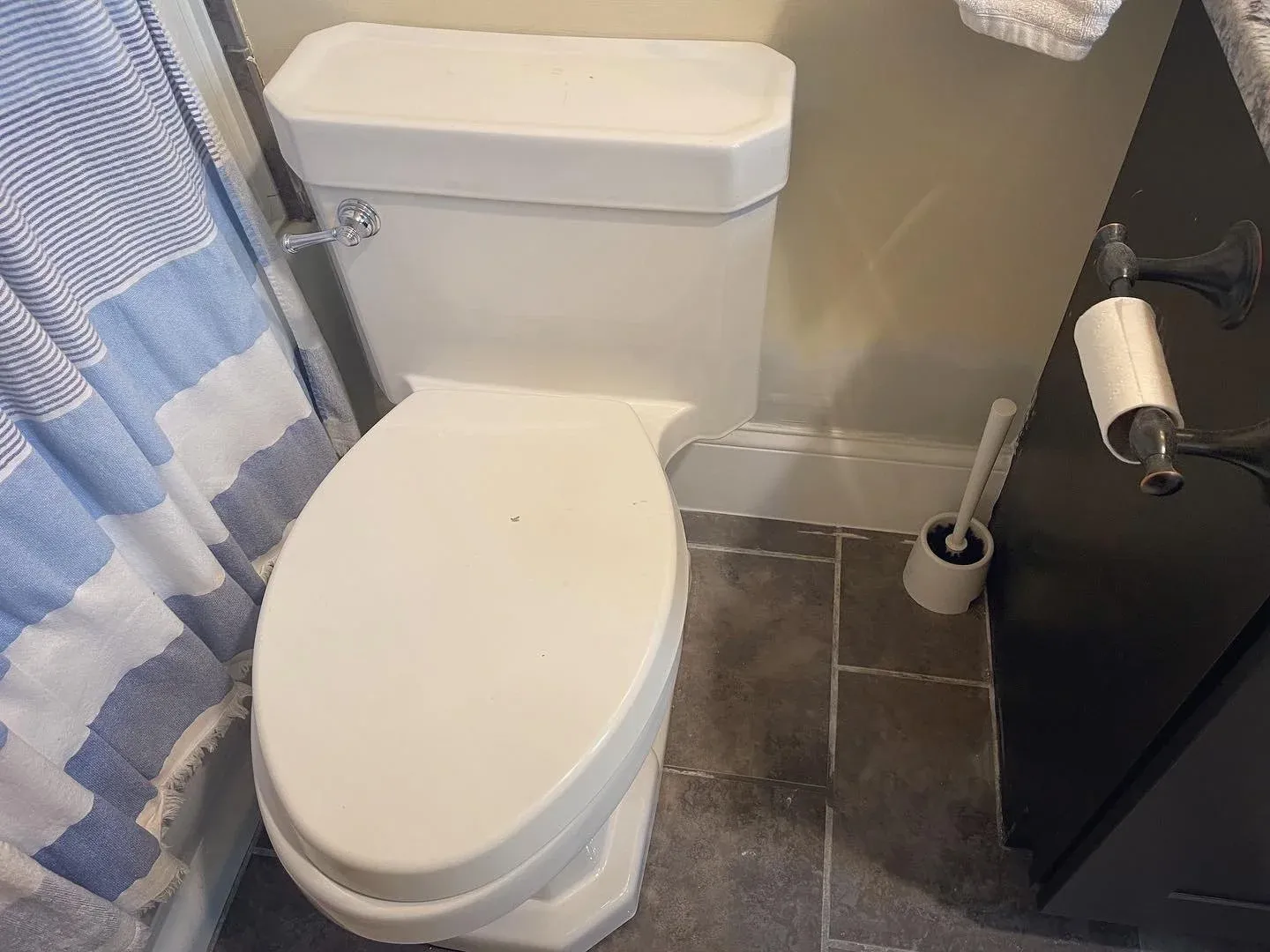 A bathroom with a white toilet, gray tiled floor, black cabinet, and a toilet paper holder with a roll of toilet paper. A toilet brush is in a holder next to the cabinet. There is a blue and white striped shower curtain partially visible.