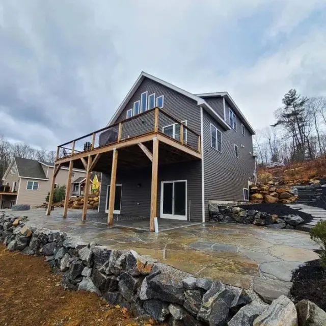 A multi-story house with dark siding and a large wooden deck overlooking a stone patio and a natural rock retaining wall.