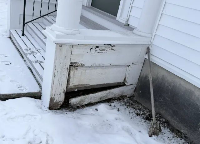 Snow-covered porch steps with a damaged white wooden skirting panel beside a house.