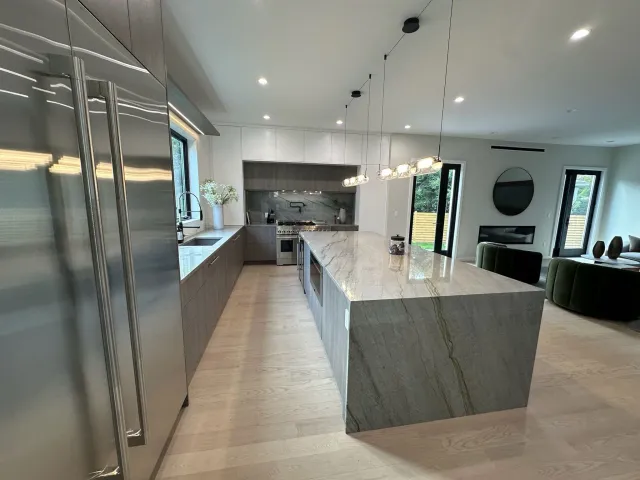 Modern kitchen with a large marble island, stainless steel appliances, and light wood flooring. There is a vase with flowers near the window and a living area with green chairs and a black round wall decor in the background.