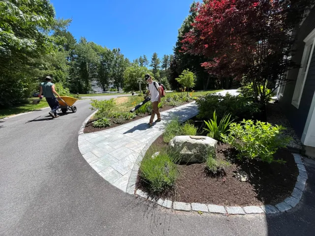 Two people working outside, one pushing a wheelbarrow and the other spraying plants with a hose on a curved stone walkway in a green, sunny yard.