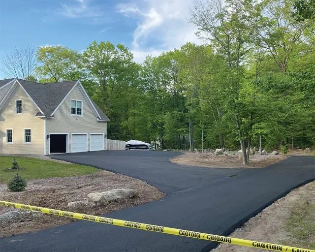 Asphalt driveway leading to a light-colored house with a three-car garage, surrounded by trees. Yellow caution tape.