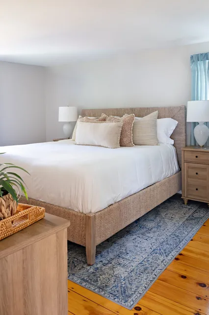 Indian Road coastal bedroom with woven bed frame, white bedding, blue area rug, and pine floors