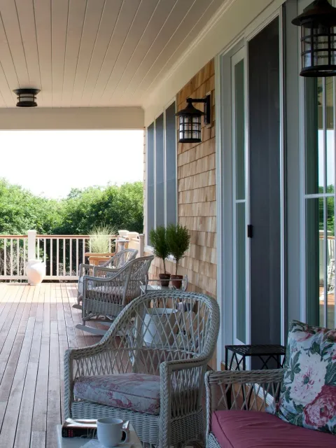 New England–style covered porch with wooden flooring, traditional wicker chairs with cushions, a small table holding a mug, potted plants, and a view of green trees.