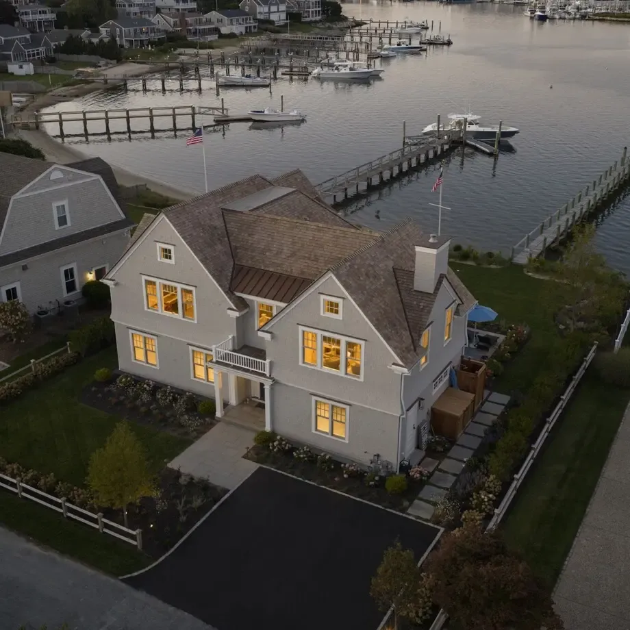Aerial view of a coastal neighborhood with a light-gray two-story house and gabled roof beside a marina, showing docks, boats, and calm water.