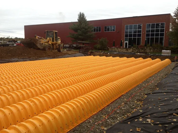 Construction site with orange drainage pipes, a bulldozer, and a brick building in the background.