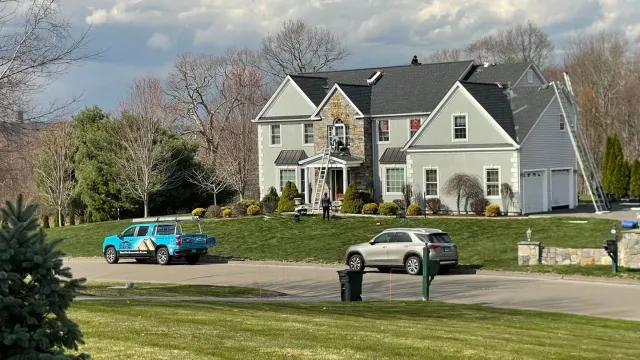 Two-story gray house with stone accents, vehicles parked on the street. Overcast sky.