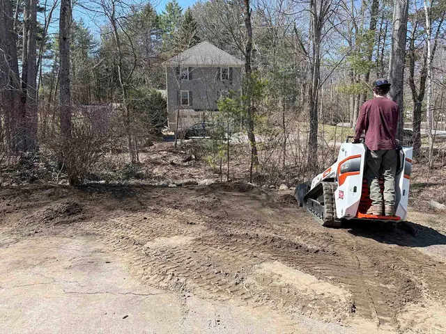 Person operating a skid steer on a dirt driveway beside a wooded lot and house under construction