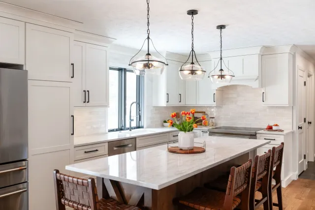 Bright white kitchen with island, pendant lights, marble counters, and a vase of orange flowers