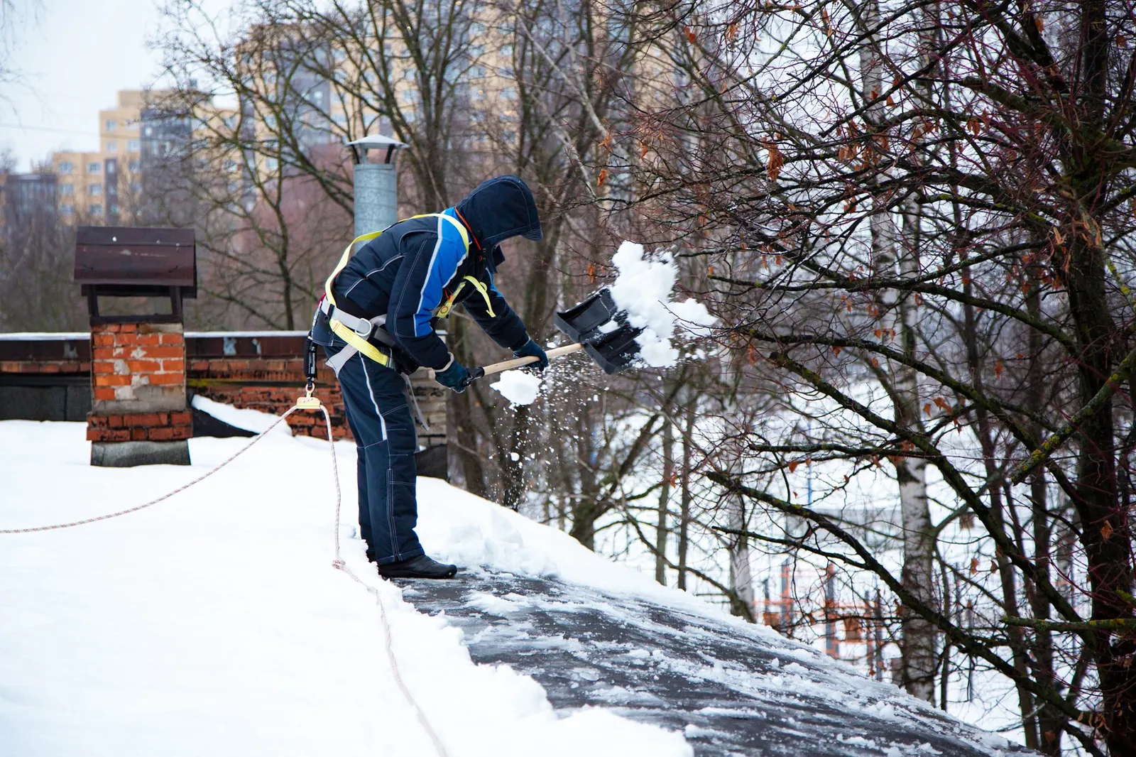 A worker in a safety harness shoveling snow from the edge of a roof