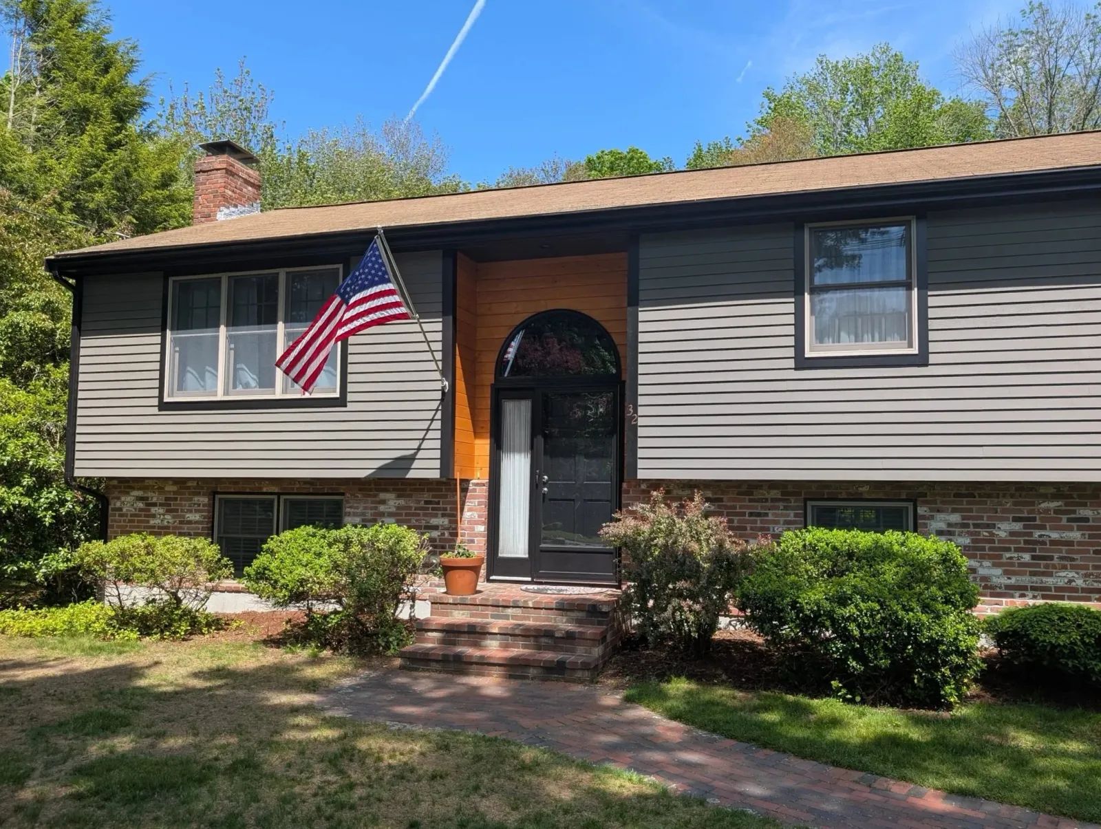 Front view of a two-story house with gray siding, brick foundation, a black front door, and an American flag in the yard, with green trees and a clear blue sky in the background.