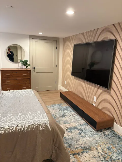Bedroom with wall-mounted TV, wood console, patterned accent wall, and wood dresser. Blue rug and beige bed.