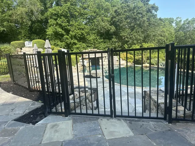 Black aluminum pool safety fence with a self-closing gate around an inground pool on a bluestone patio with stone waterfall