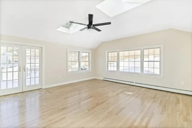 Bright, empty room with light hardwood floors, white walls, large windows, a ceiling fan, and a glass door to the outside.
