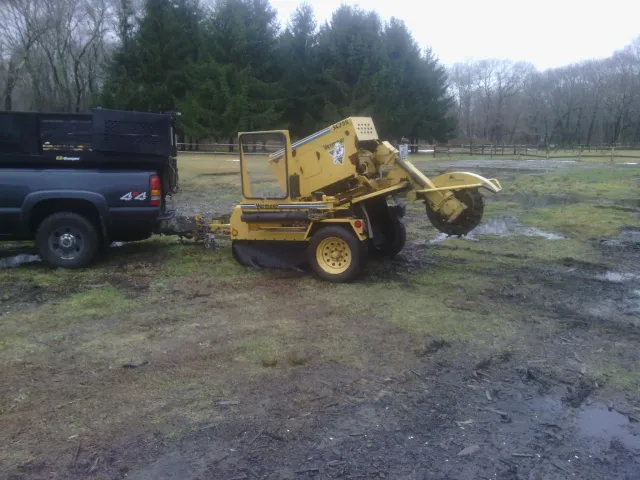 A yellow Vermeer stump grinder attached to a black pickup truck in a grassy outdoor area with trees and a wooden fence in the background.
