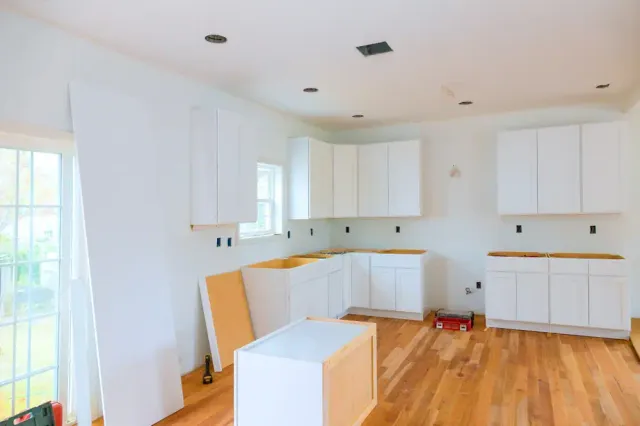 A kitchen under renovation in MA features white cabinets being installed, some doors leaning against the wall, and tools scattered on a wood floor. No appliances or countertops are present yet—a glimpse of Home Remodeling Essex County in progress.