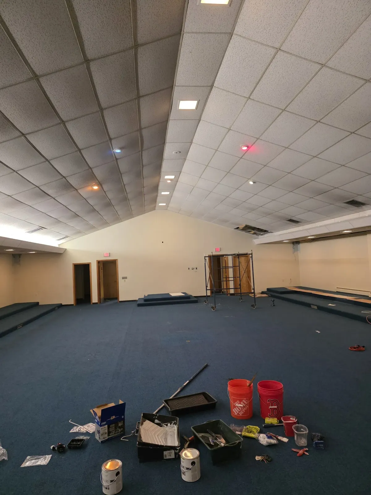 Large empty room with blue carpet, white ceiling tiles, and scattered tools and supplies on the floor.