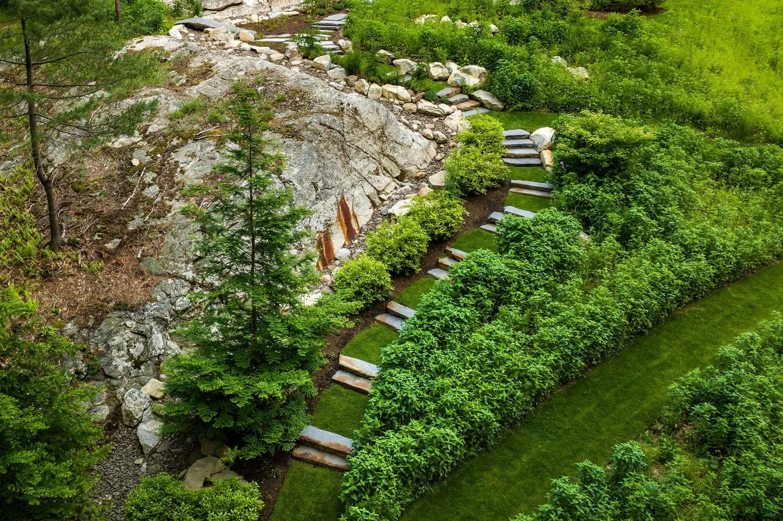 This image displays a lush garden with stepping stones, manicured hedges, and a rocky outcrop surrounded by vibrant greenery from an aerial perspective.