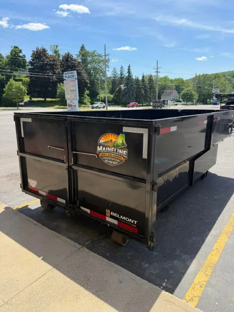 Black construction dumpster in a parking lot under a blue sky, with trees and road signs behind it